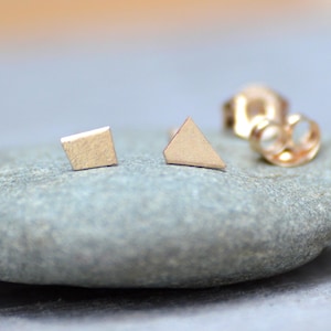 May include: Two minimalist gold earrings, one shaped like a square and the other like a triangle, on a gray stone background.