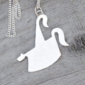 May include: A silver-colored pendant necklace featuring a stylized, abstract design. The pendant has a unique shape with curved elements and is suspended from a delicate chain. The necklace is displayed on a wooden surface.