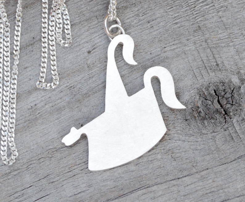 May include: A silver-colored pendant necklace featuring a stylized, abstract design. The pendant has a unique shape with curved elements and is suspended from a delicate chain. The necklace is displayed on a wooden surface.