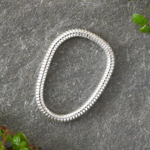 Könnte beinhalten: Ein silbernes Schlangenkettenarmband, hergestellt aus kleinen, ineinandergreifenden Gliedern. Das Armband liegt flach auf einer grauen, strukturierten Oberfläche.