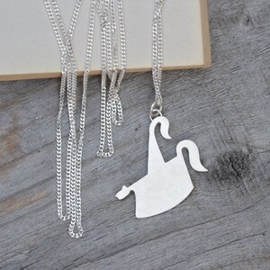 May include: A silver-colored necklace with a pendant shaped like a stylized ghost. The pendant hangs from a delicate chain, resting on a wooden surface. The ghost design features curved lines and a simple, elegant form.