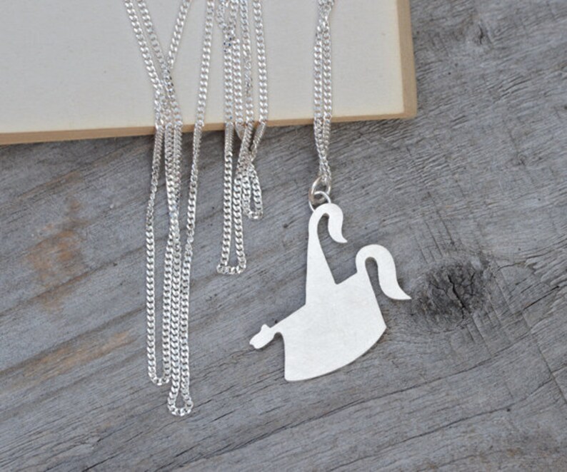 May include: A silver-colored necklace with a pendant shaped like a stylized ghost. The pendant hangs from a delicate chain, resting on a wooden surface. The ghost design features curved lines and a simple, elegant form.