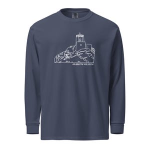 May include: A navy blue long-sleeve shirt featuring a white line drawing of a lighthouse on a rocky landscape. The coordinates 43.8661°N 103.5323°W are printed below the image. The shirt has a crew neck and long sleeves.