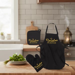 May include: A black kitchen set featuring an apron, oven mitt, and pot holder. Each item has gold text that reads "Ramadan Mubarak" with a crescent moon and star design. The set is displayed on a wooden countertop.