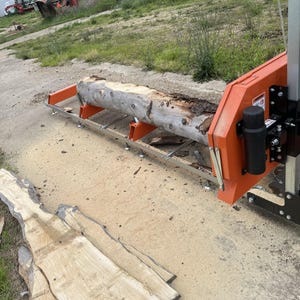 May include: A close-up of a wood-cutting machine with a large log on the bed. The machine has a metal frame and a red and black motor. A wooden plank is on the ground in front of the machine.