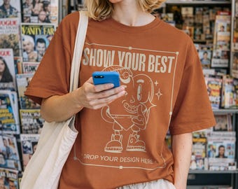 May include: A rust-colored t-shirt with the text "SHOW YOUR BEST" and a cartoon character design. The shirt is paired with light-colored shorts. The background features a magazine stand.