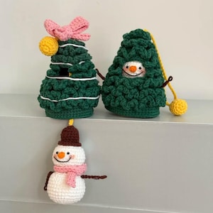 May include: Crocheted Christmas ornaments. Two green Christmas trees with white accents, one with a pink bow and yellow ball, the other with a snowman face. A crocheted snowman with a brown hat, pink scarf, and stick arms.