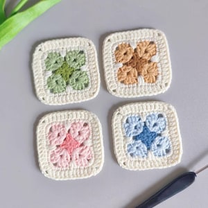 May include: Four handmade crochet coasters in a square shape. Each coaster features a different color scheme: green, brown, pink, and blue, with a white border. A crochet hook is visible in the bottom right corner. A pink tulip is in the top left corner.