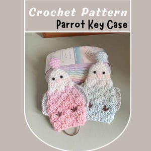 May include: Two crocheted parrot keychains, one pink and one blue, with black eyes and brown beaks. The keychains are hanging from a silver key ring.