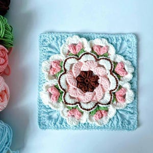 May include: A crocheted square with a large pink and white flower in the center. The flower has a brown center and green leaves. The square is surrounded by a blue border.