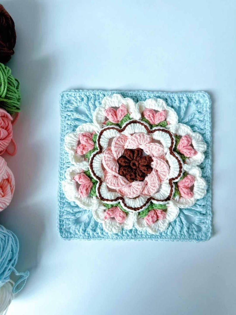 May include: A crocheted square with a large pink and white flower in the center. The flower has a brown center and green leaves. The square is surrounded by a blue border.
