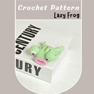 Può includere: Una rana verde all'uncinetto con una lingua rosa sdraiata su un libro bianco con la parola "CENTURY" stampata sulla copertina. Il libro è intitolato "Lazy Frog" in testo nero.