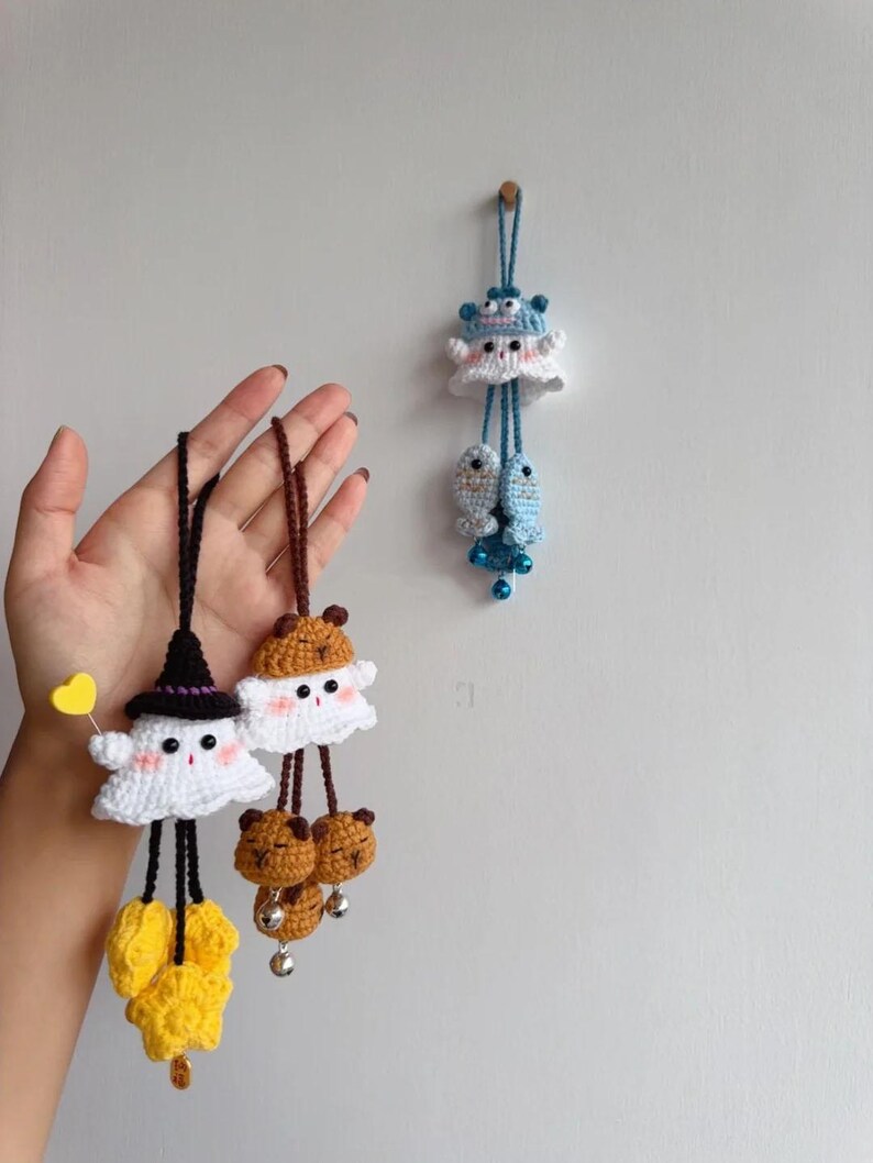 May include: Crocheted ghost and bear-themed hanging ornaments in white, black, brown, and yellow. One ghost wears a witch hat and holds a yellow heart. The other has bear ears. A blue sea creature ornament hangs on the wall.