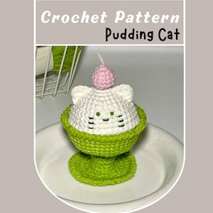 May include: A crocheted pudding cat amigurumi toy with a white body, green bowl, and pink cherry on top. The toy is sitting on a white plate.