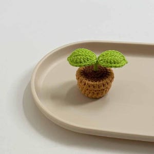 May include: A crocheted plant in a small brown pot with two green leaves. The plant is sitting on a beige oval tray. The plant is a handmade item.