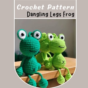 May include: Two green crocheted frogs with white eyes and smiling mouths. The frogs have long dangling legs and are sitting on a wooden surface. Crochet pattern Dangling Legs Frog.
