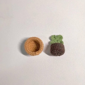 May include: Two crocheted items on a white surface. One is a small, open-topped, tan-colored bowl. The other is a miniature plant with a brown pot and green leaves, all crocheted. The items are likely handmade.