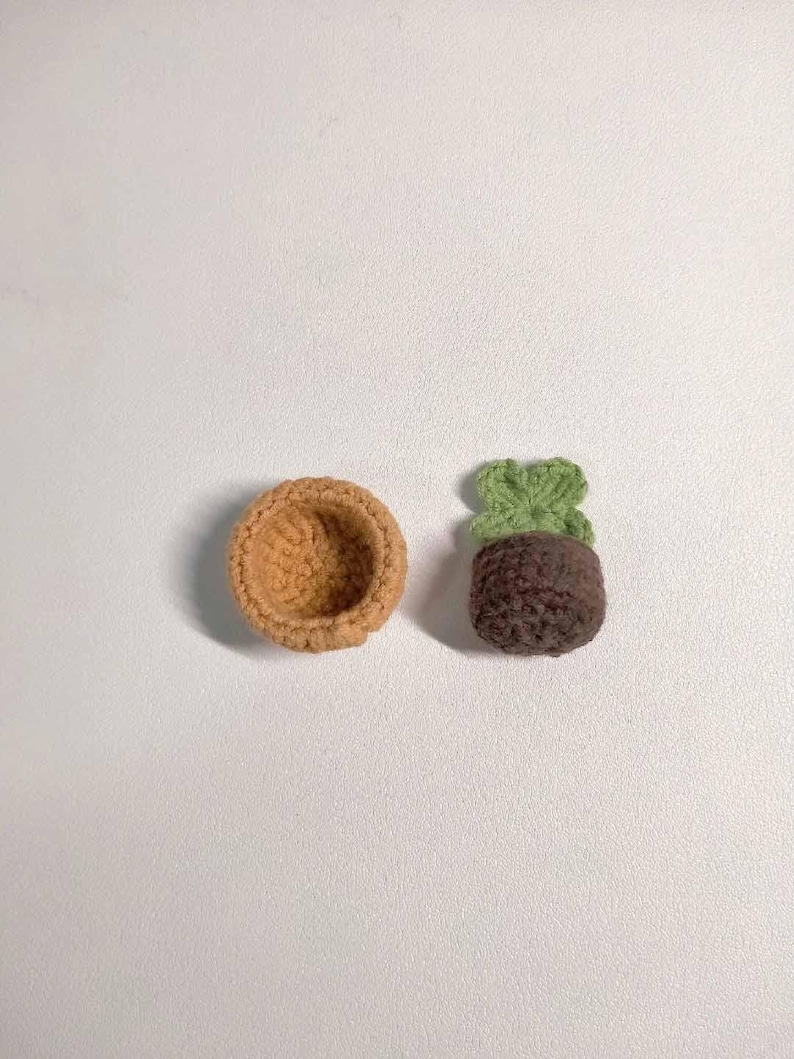 May include: Two crocheted items on a white surface. One is a small, open-topped, tan-colored bowl. The other is a miniature plant with a brown pot and green leaves, all crocheted. The items are likely handmade.