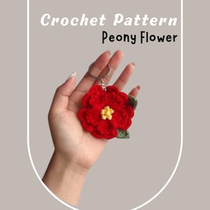May include: A red crocheted peony flower with a yellow center and green leaves attached to a silver key chain.