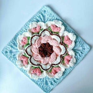 May include: A crocheted square with a large, multi-colored flower in the center. The flower is made of pink, white, green, and brown yarn. The square is made of light blue yarn.