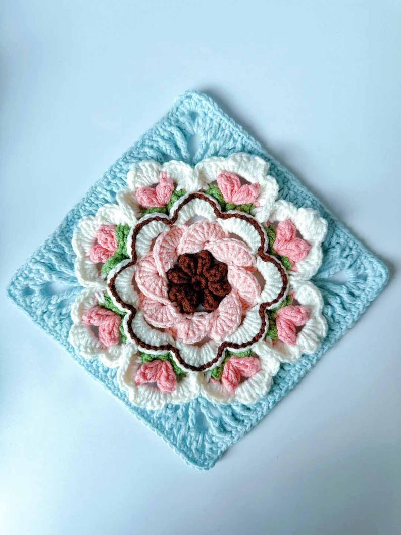 May include: A crocheted square with a large, multi-colored flower in the center. The flower is made of pink, white, green, and brown yarn. The square is made of light blue yarn.