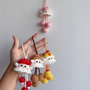May include: Handmade crochet mushroom and ghost-shaped ornaments in white, red, brown, and yellow. The ornaments have dangling mushroom or ghost-shaped accents. One ornament is hanging on a wall. The other ornaments are held in a hand.
