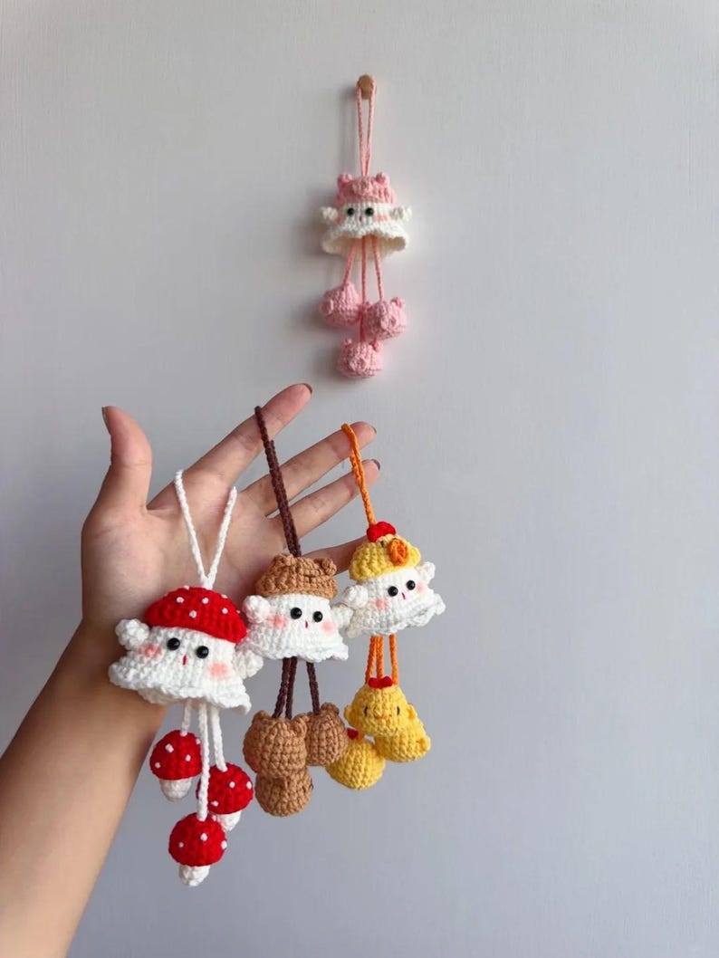 May include: Handmade crochet mushroom and ghost-shaped ornaments in white, red, brown, and yellow. The ornaments have dangling mushroom or ghost-shaped accents. One ornament is hanging on a wall. The other ornaments are held in a hand.