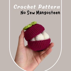 May include: A crocheted mangosteen fruit with a green top and a white interior. The fruit is open to reveal the white interior. The fruit is held in a hand.