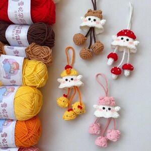 May include: Crocheted mushroom and animal-themed keychains in white, brown, red, and pink. The keychains have dangling elements and are displayed with colorful yarn skeins in the background.