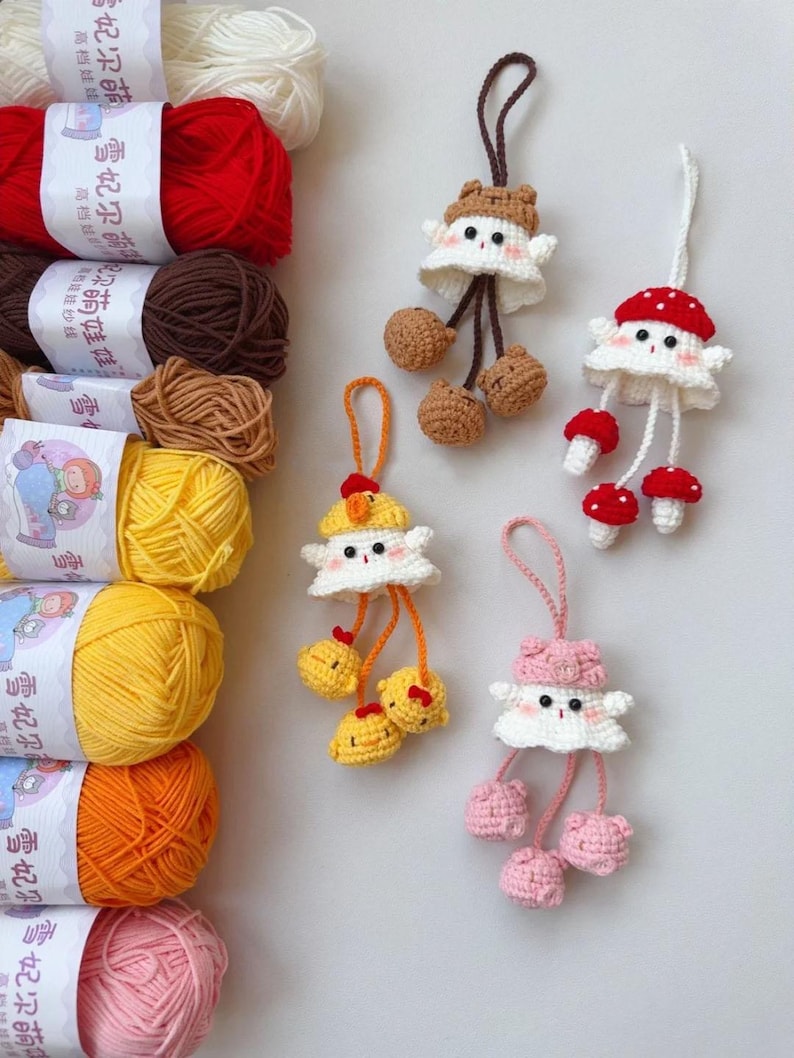 May include: Crocheted mushroom and animal-themed keychains in white, brown, red, and pink. The keychains have dangling elements and are displayed with colorful yarn skeins in the background.