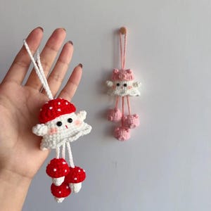 May include: Crocheted mushroom-themed ornaments. One ornament features a red and white mushroom cap with a smiling face and three red and white mushroom charms. The other ornament is pink and white with a mushroom cap design and three pink pom-poms.