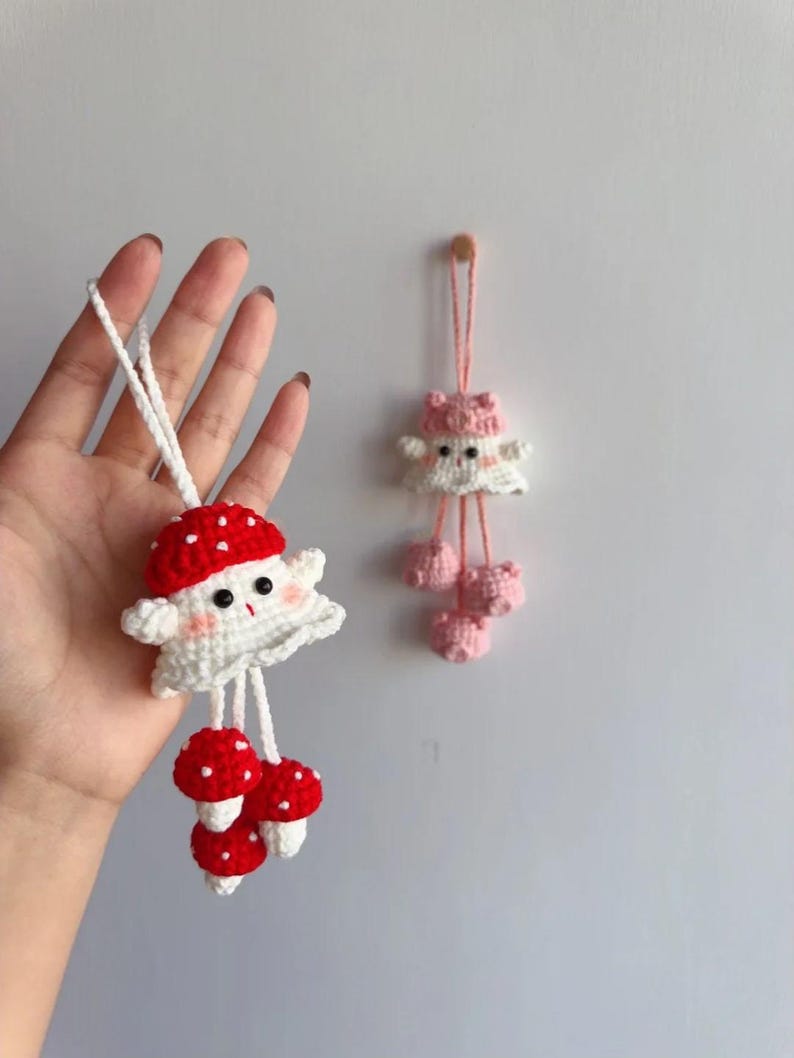 May include: Crocheted mushroom-themed ornaments. One ornament features a red and white mushroom cap with a smiling face and three red and white mushroom charms. The other ornament is pink and white with a mushroom cap design and three pink pom-poms.