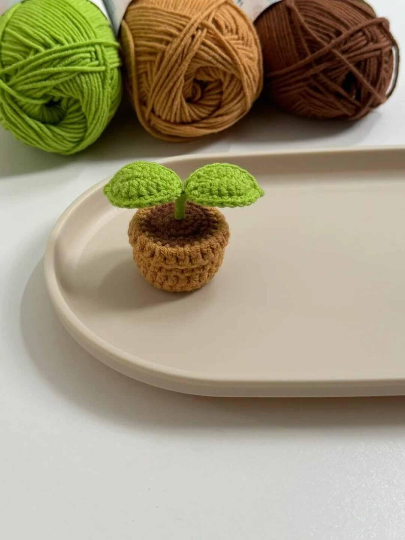May include: A crocheted miniature plant with two green leaves in a brown pot, set on a beige oval tray. Three balls of yarn in green, tan, and brown are in the background. The handmade plant is a charming decorative item.