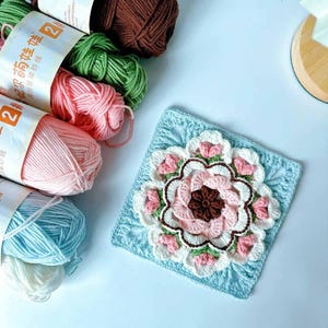 May include: A crocheted square with a large pink flower in the center. The flower has a brown center and is surrounded by smaller pink flowers. The square is on a light blue background. There are several balls of yarn in different colors in the background.