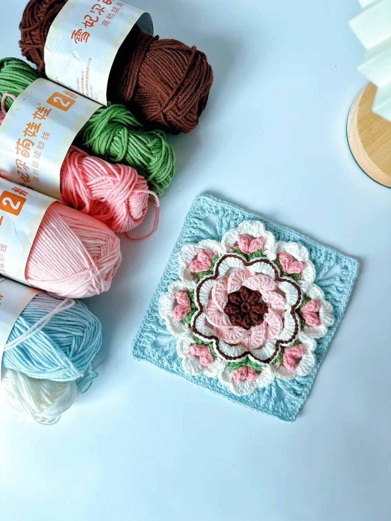 May include: A crocheted square with a large pink flower in the center. The flower has a brown center and is surrounded by smaller pink flowers. The square is on a light blue background. There are several balls of yarn in different colors in the background.