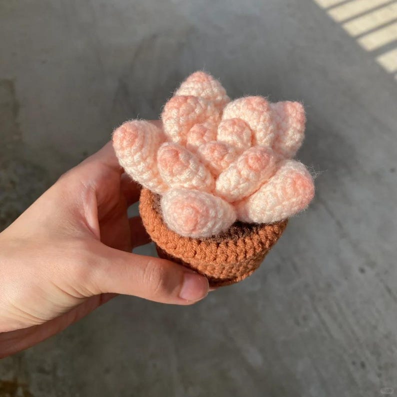 May include: A crocheted succulent plant in a brown pot. The succulent is pink and white and has a realistic appearance.