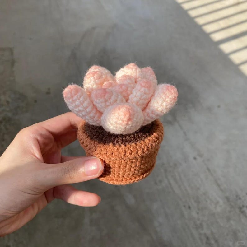 May include: A small crocheted succulent plant in a brown pot. The succulent is pink and white and has a cluster of leaves.