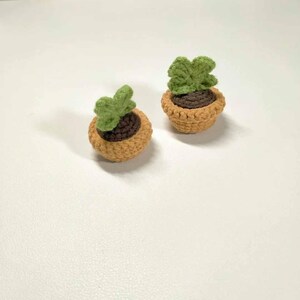 May include: Two miniature crochet potted plants. Each plant features a brown pot with a light green plant. The pots are a light brown color. The plants are on a white surface.