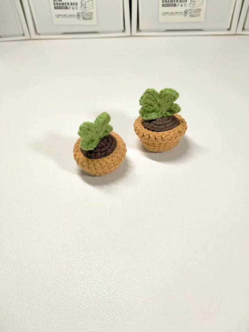 May include: Two miniature crochet potted plants. Each plant features a brown pot with a light green plant. The pots are a light brown color. The plants are on a white surface.