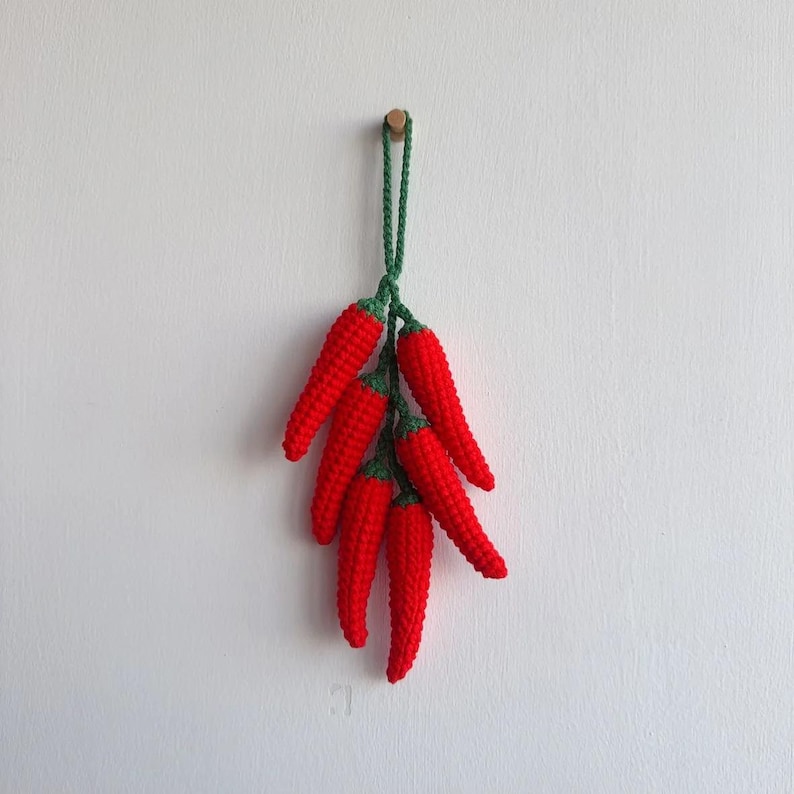 May include: A bunch of red crocheted chili peppers hanging from a string. The peppers are green at the top and red at the bottom.
