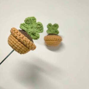 May include: Two crochet succulent plants. One is attached to a metal rod. The pots are tan and the plants are green. The soil is brown. The plants are in the shape of a four-leaf clover.