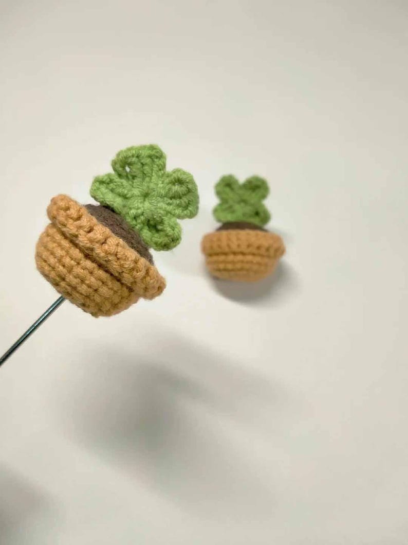 May include: Two crochet succulent plants. One is attached to a metal rod. The pots are tan and the plants are green. The soil is brown. The plants are in the shape of a four-leaf clover.