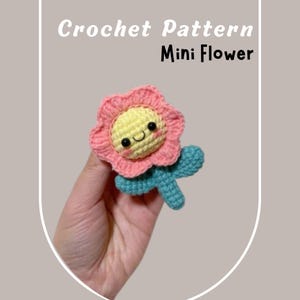 Puede incluir: Una pequeña flor de crochet con una flor rosa y un centro amarillo con una cara sonriente. La flor tiene un tallo y hojas azules.