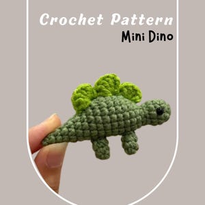 May include: Crochet pattern for a mini green dinosaur with a green spiky back. The dinosaur is being held by a hand.