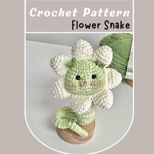 May include: A crocheted flower snake amigurumi toy with a green body and white flower petals. The toy has a cute face with black eyes and a pink blush. The toy is sitting on a wooden base.