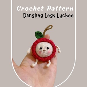 Può includere: Un modello all'uncinetto di un frutto di litchi con gambe pendenti. Il frutto ha un anello esterno rosso, un centro bianco, una foglia verde e un gambo marrone. Il testo "Crochet Pattern Dangling Legs Lychee" è nella parte superiore dell'immagine.