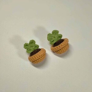 May include: Two crocheted plant pot earrings. Each earring features a small, light brown pot with dark brown 'soil' and a green, four-leaf clover-shaped plant. The earrings are handmade and likely suitable for everyday wear.
