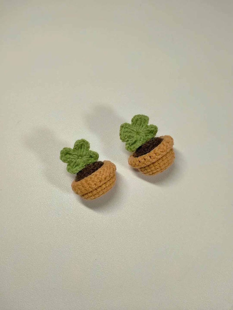May include: Two crocheted plant pot earrings. Each earring features a small, light brown pot with dark brown 'soil' and a green, four-leaf clover-shaped plant. The earrings are handmade and likely suitable for everyday wear.