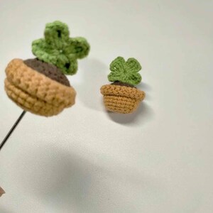 May include: Two crocheted succulent plants. One is attached to a black wire. The plants have green leaves and are potted in brown and tan crocheted pots. The background is a plain white surface.