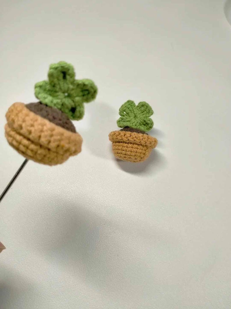 May include: Two crocheted succulent plants. One is attached to a black wire. The plants have green leaves and are potted in brown and tan crocheted pots. The background is a plain white surface.